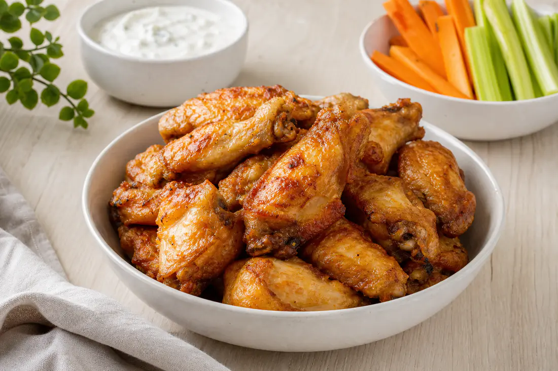 Alitas de pollo doradas y crujientes apiladas en un cuenco, con salsa para mojar y palitos de apio