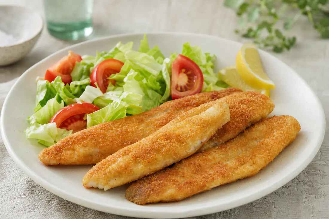 Filetes de bacaladilla rebozados y dorados sobre un plato con ensalada de lechuga y una cuña de limón