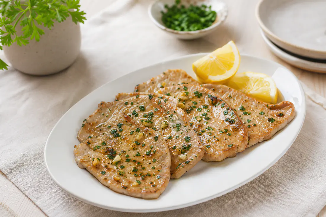 Cuatro filetes de ternera finos y dorados con ajo y perejil picado, servidos en un plato con rodajas de limón