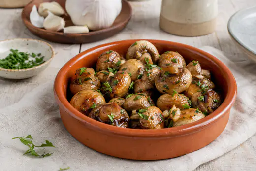 Champiñones enteros dorados con ajo y perejil picado, servidos en una cazuelita de barro sobre una mesa rústica