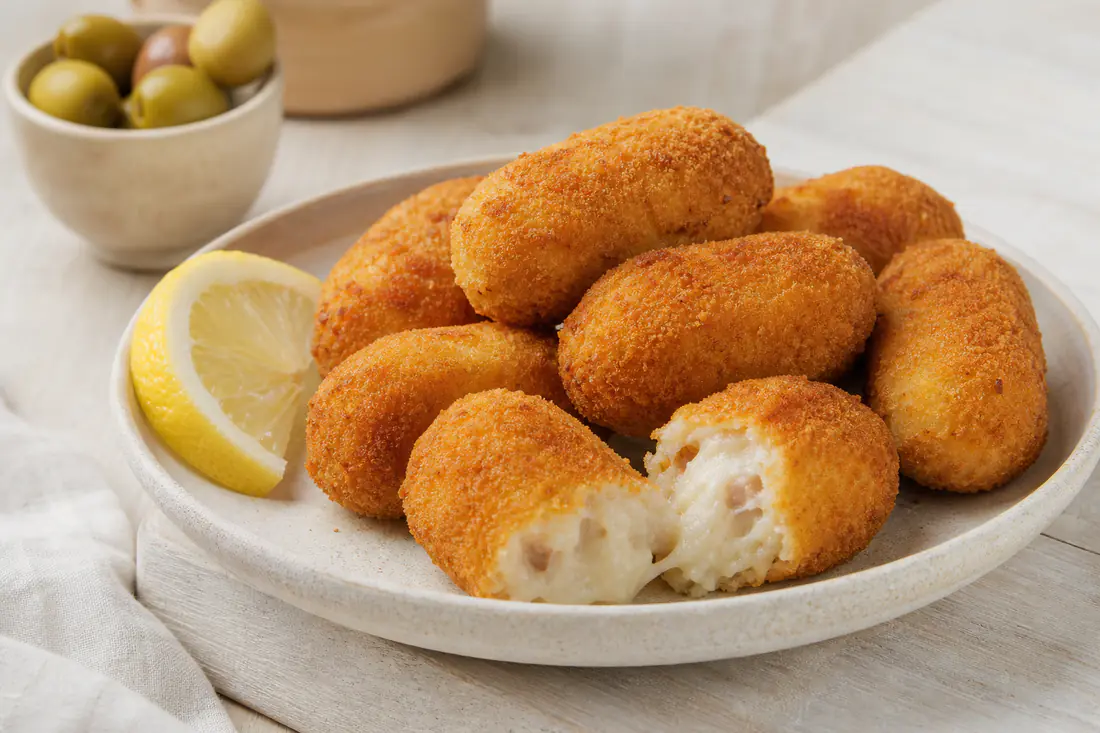 Croquetas doradas y crujientes en un plato blanco, una partida por la mitad mostrando la bechamel cremosa, con limón