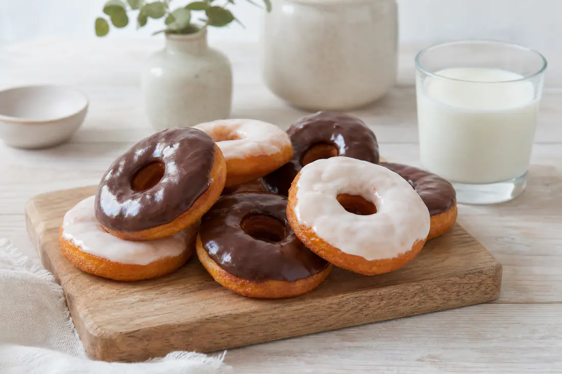 Donuts caseros con glaseado blanco y de chocolate sobre una tabla de madera, con un vaso de leche al lado
