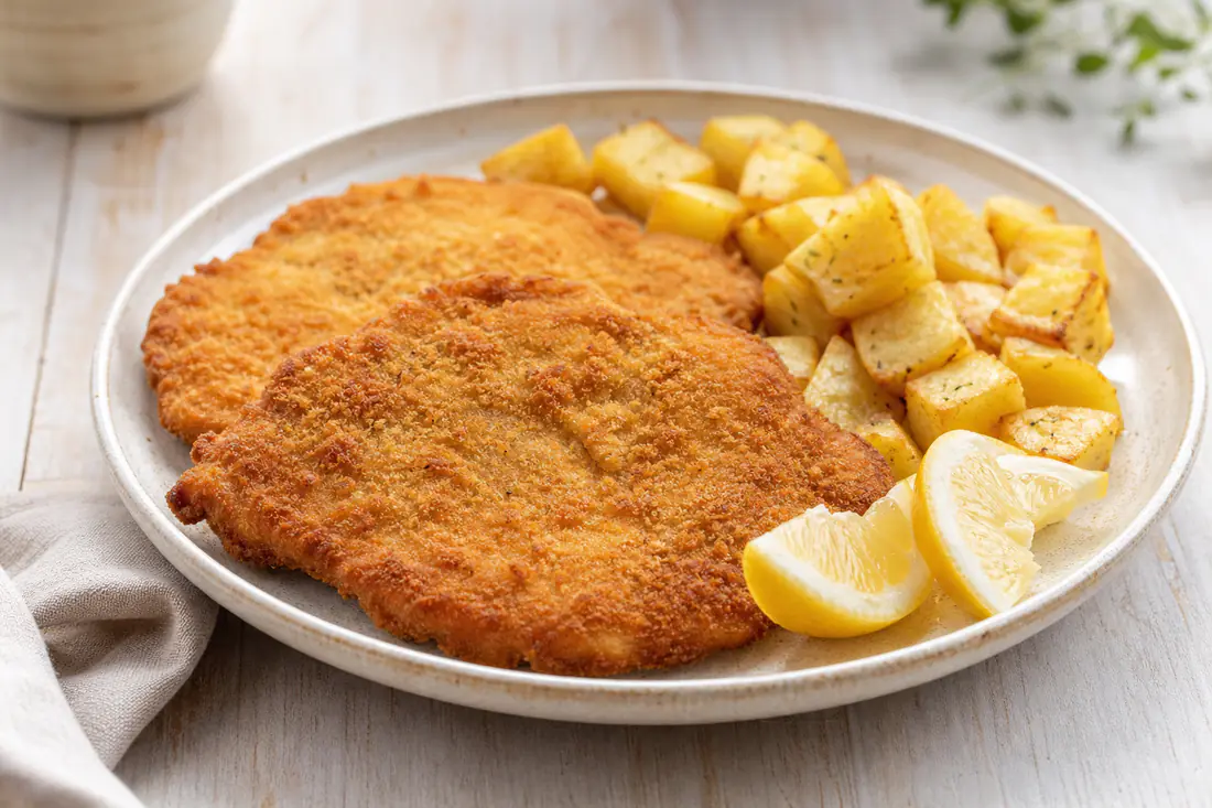 Dos escalopes de ternera empanados, dorados y crujientes, servidos con rodajas de limón y patatas