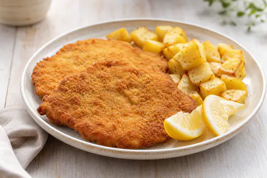Dos escalopes de ternera empanados, dorados y crujientes, servidos con rodajas de limón y patatas