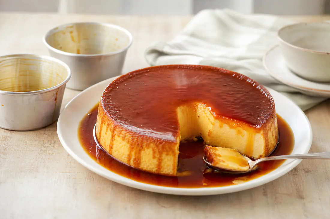 Flan de huevo desmoldado en un plato blanco, con el caramelo oscuro cayendo por los lados, junto a las flaneras vacías