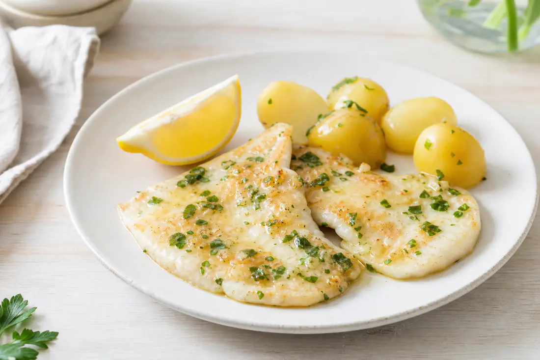 Dos filetes de gallo dorados con mantequilla de ajo, perejil y una cuña de limón sobre un plato blanco