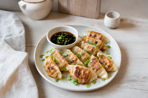 Gyozas doradas y crujientes en un plato, con un cuenco de salsa de soja y vinagre y cebolleta picada al lado