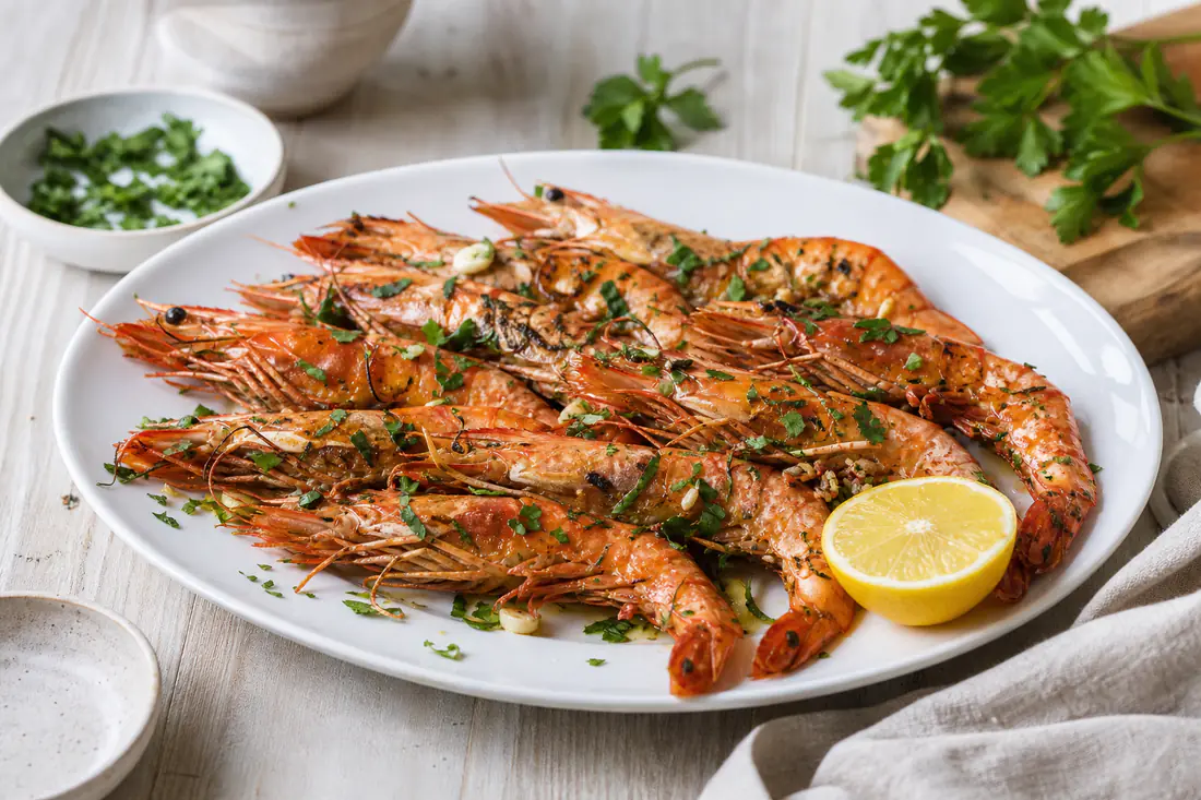 Langostinos con cáscara asados, con ajo y perejil picado, sobre una fuente blanca con medio limón