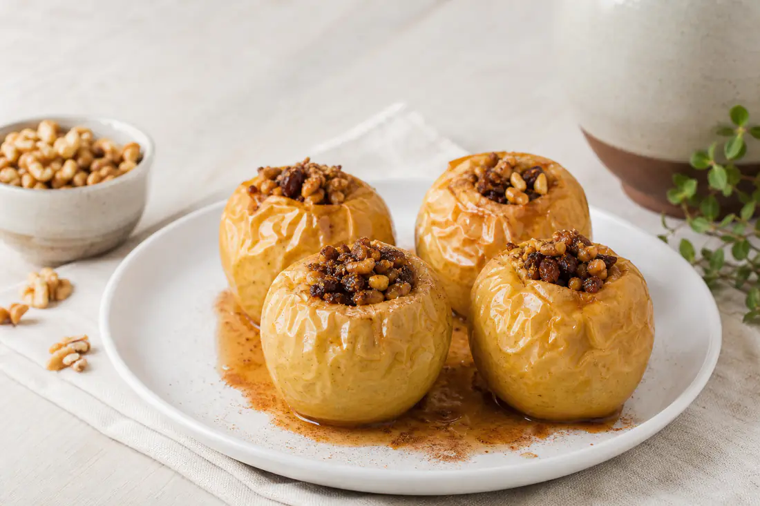 Cuatro manzanas asadas rellenas de nueces y pasas, con la piel arrugada y brillante de miel, servidas en un plato con canela