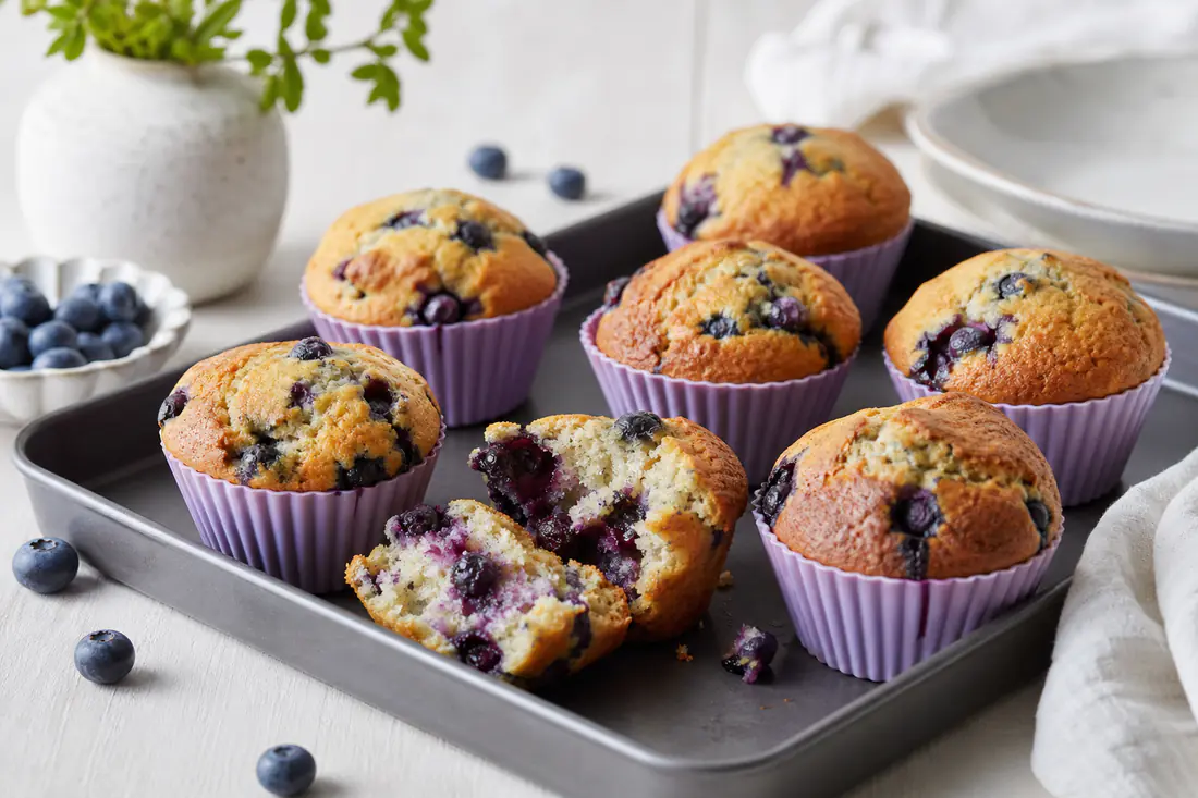 Muffins de arándanos con la parte superior dorada y arándanos visibles, en cápsulas de silicona sobre una bandeja