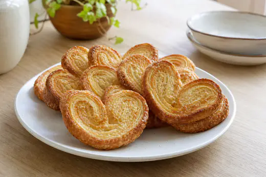 Palmeritas de hojaldre doradas y caramelizadas, con forma de corazón, dispuestas en un plato blanco sobre una mesa de madera