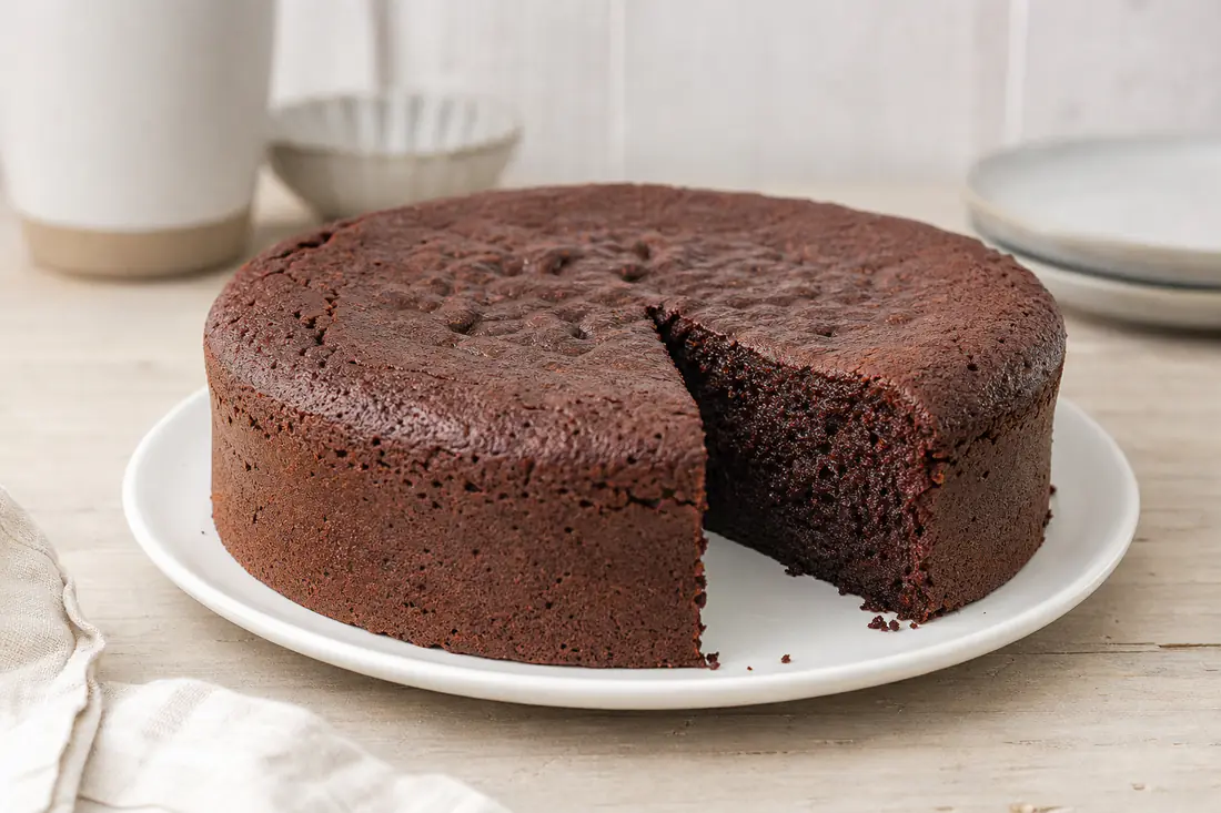 Bizcocho de chocolate redondo y esponjoso con una porción cortada que muestra la miga húmeda, sobre un plato blanco