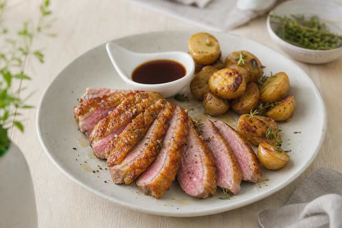 Pechuga de pato cortada en lonchas, con piel dorada y centro rosado, sobre un plato con patatas y salsa