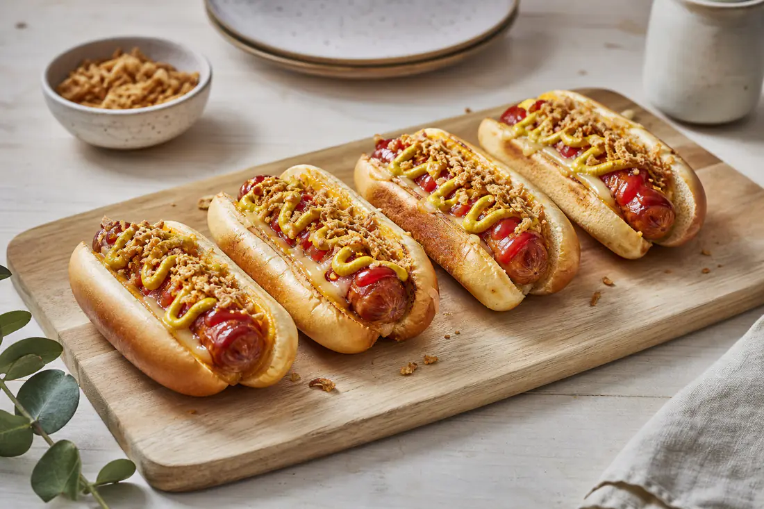 Cuatro perritos calientes con salchicha dorada y queso fundido en pan tostado, con mostaza, ketchup y cebolla frita crujiente