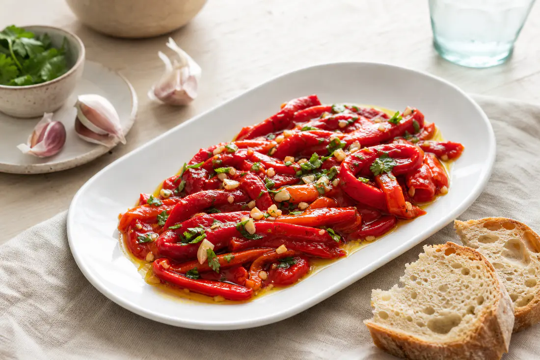 Pimientos rojos asados y pelados en tiras, con ajo y perejil, sobre una fuente blanca con un chorrito de aceite de oliva