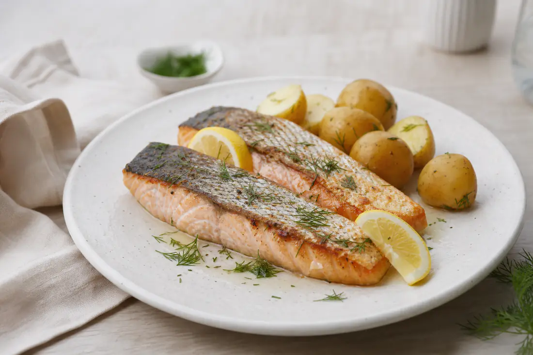 Dos filetes de salmón con la piel crujiente y la carne rosada, con eneldo fresco y limón sobre un plato blanco con patatas baby