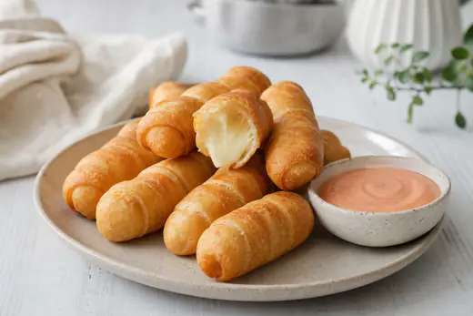 Tequeños dorados y crujientes, uno partido por la mitad con el queso fundido, servidos con un cuenco de salsa rosa