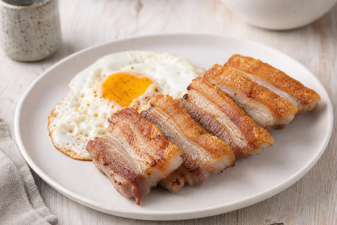 Lonchas de panceta fresca doradas y crujientes, con un borde de grasa tostada, servidas en un plato blanco